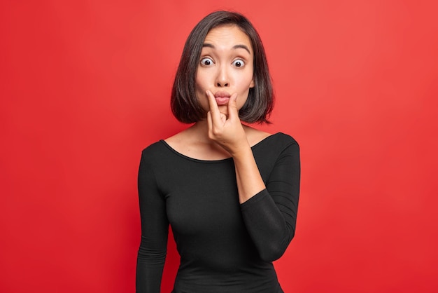 Surprised brunette young woman puckers lips at camera looks surprisingly wears black dress has slim figure poses against vivid red background looks shocked stands indoor. Human facial expressions