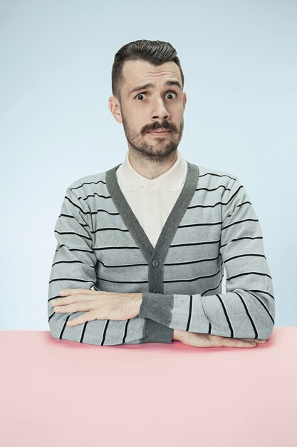 Удивленный деловой человек, сидящий за столом на синем фоне студии. Портрет в стиле минимализм