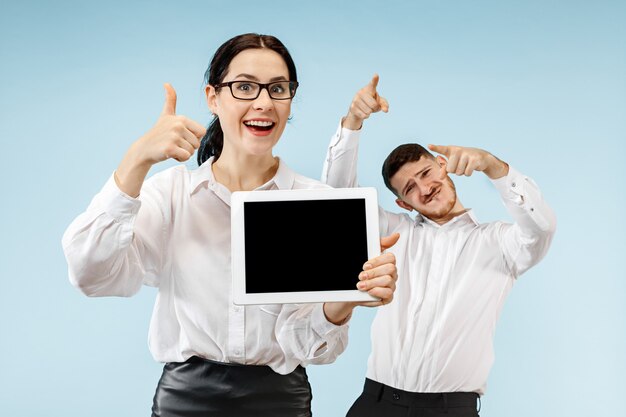 The surprised business man and woman smiling on a blue studio background and showing empty screen of laptop or tablet