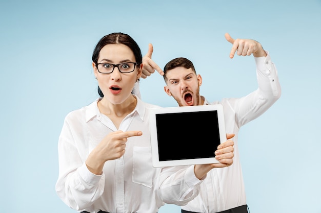 The surprised business man and woman smiling on a blue studio background and showing empty screen of laptop or tablet