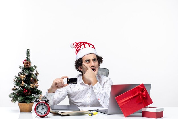 Удивленный деловой человек в шляпе санта-клауса и держит свою банковскую карту, внимательно глядя на что-то в офисе на белом фоне