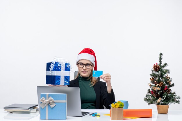 サンタクロースの帽子と白い背景の上のクリスマスの贈り物と銀行カードを保持しているテーブルに座って眼鏡をかけて驚いたビジネス女性