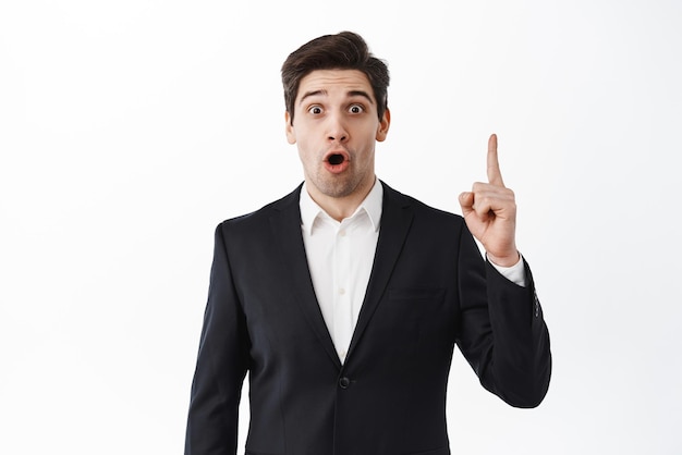 Surprised businessman pointing finger up and gasping amazed say wow stare excited at camera standing in black suit against white studio background