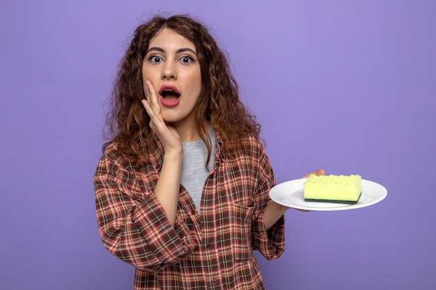 Free photo surprised calling someone young cleaning woman holding cleaning sponge on plate