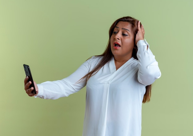 Surprised casual caucasian middle-aged woman holding and looking at phone and grabbed head isolated on olive green wall