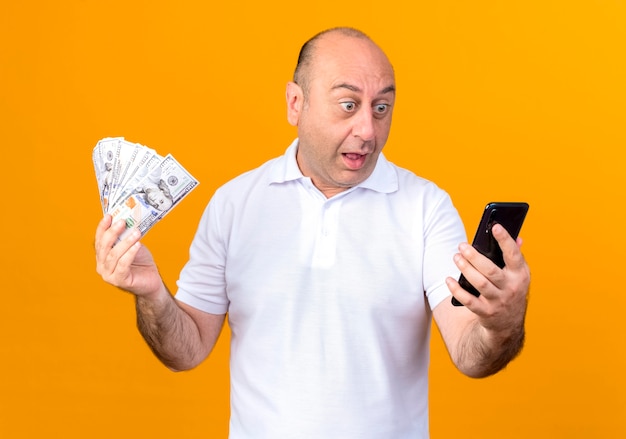Удивленный случайный зрелый мужчина держит деньги и смотрит на телефон, изолированный на желтой стене