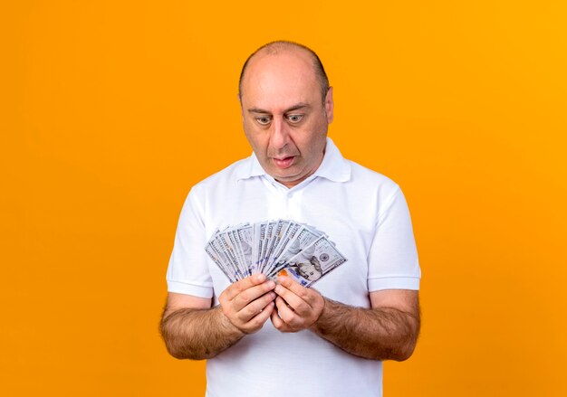 Surprised casual mature man holding and looking at cash isolated on yellow