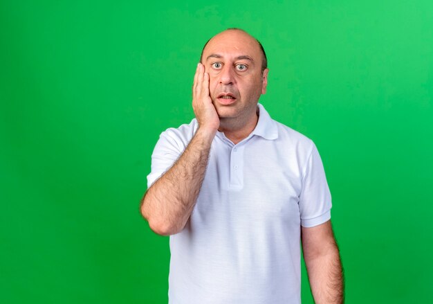 Surprised casual mature man putting hand on cheek isolated on green wall