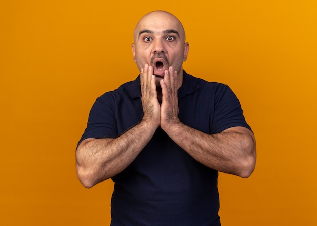 Free photo surprised casual middle-aged man keeping hands on chin looking at front isolated on orange wall