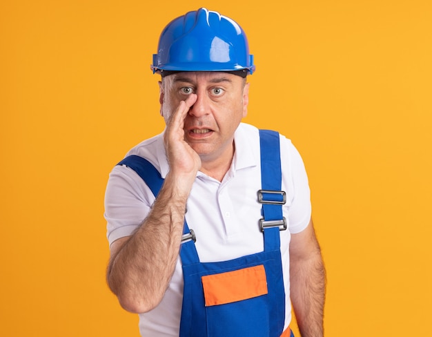 Foto gratuita l'uomo adulto caucasico sorpreso del costruttore in uniforme tiene la mano vicino alla bocca sull'arancio