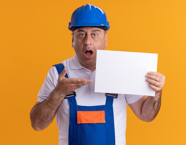 Surprised caucasian adult builder man in uniform holds and points at blank paper sheet on orange