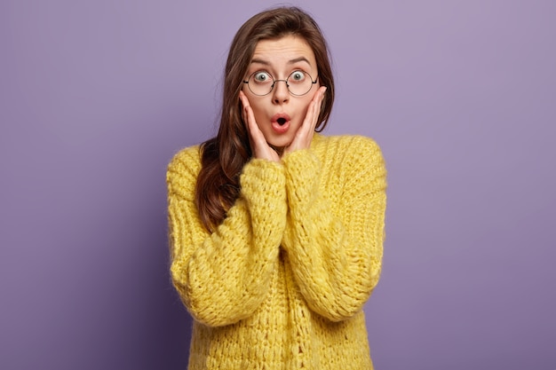 Surprised Caucasian female model keeps palms near face, expresses amazement, has mouth wide opened, can not believe in shocking news, dressed in yellow knitted jumper, isolated over purple wall