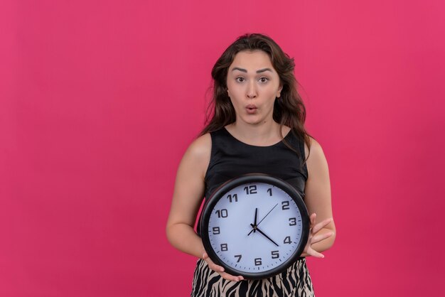 Surprised caucasian girl wearing black undershirt holding a wall clock on pink background