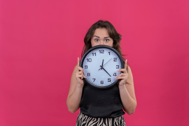 Ragazza caucasica sorpresa che porta maglietta nera che tiene un orologio da parete su fondo rosa