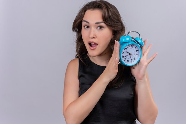 Surprised caucasian young girl wearing black undershirt holding a alarm clock on white background