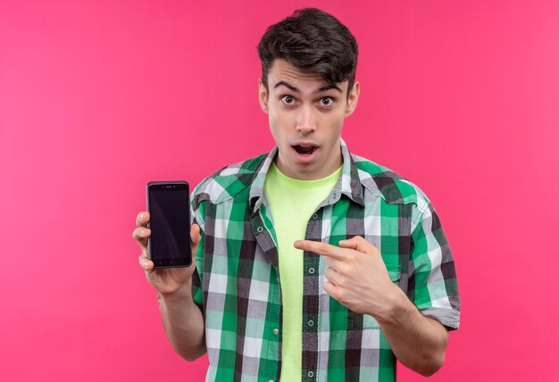 Surprised caucasian young guy wearing green shirt holding and points to phone on isolated pink background