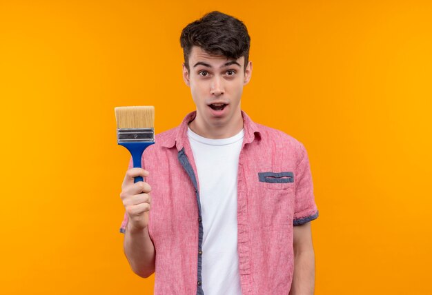 Surprised caucasian young guy wearing pink shirt holding paint brush on isolated orange background