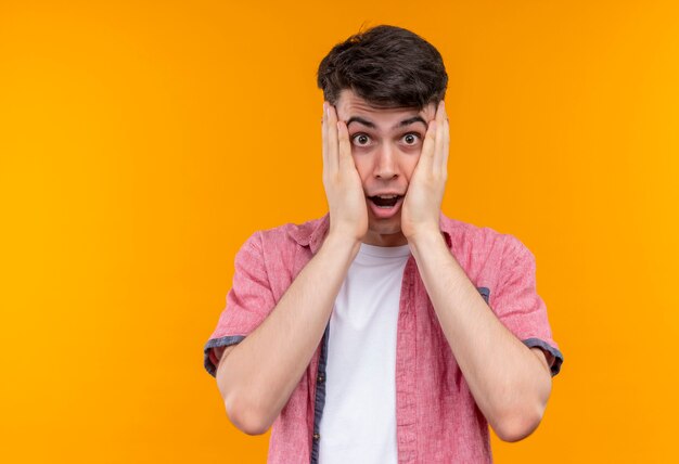 Surprised caucasian young guy wearing pink shirt put his hands on cheeks on isolated orange background