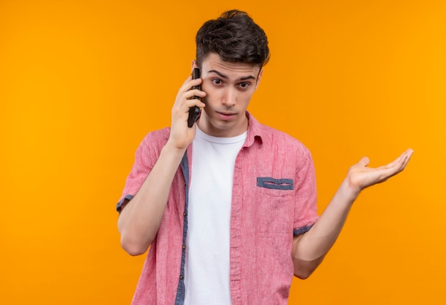 Surprised caucasian young guy wearing pink shirt speaks on phone raised hand on isolated orange background