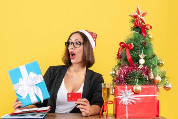 산타 클로스 모자와 노란색에 사무실에서 선물 및 은행 카드를 보여주는 안경 정장에 놀란 매력적인 아가씨