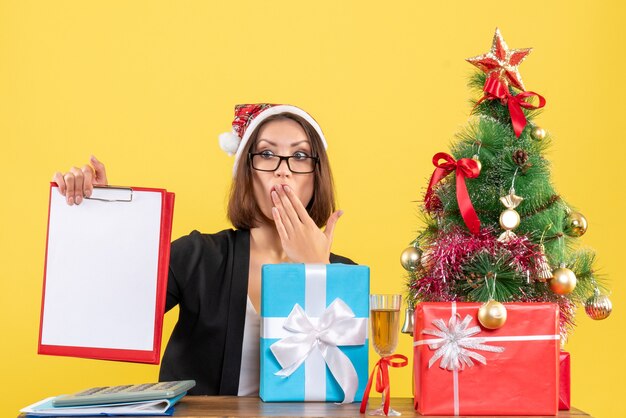 Surprised charming lady in suit with santa claus hat looking at document in the office on yellow isolated 