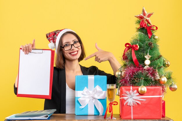 Surprised charming lady in suit with santa claus hat looking at document pointing herself in the office on yellow isolated 