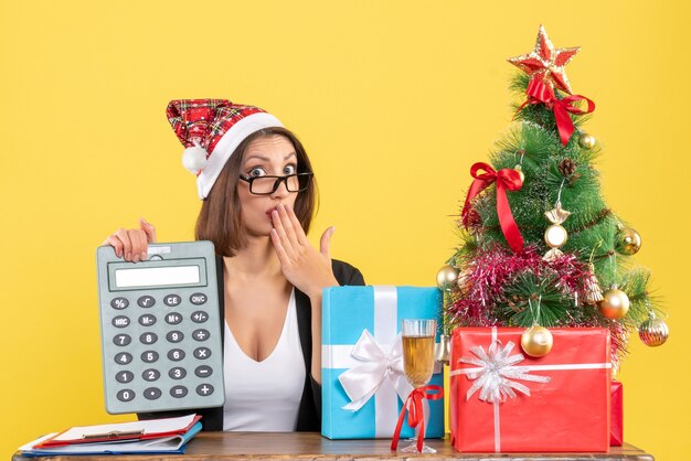 Surprised charming lady in suit with santa claus hat showing calculator in the office on yellow isolated 