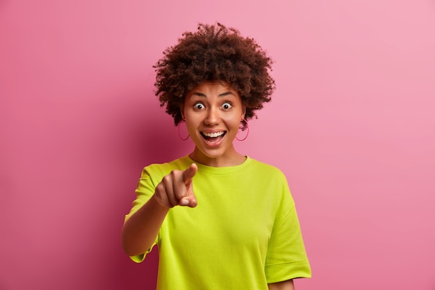 Surprised cheerful afro american woman dressed in casual clothes points directly at you chooses something has shocked glad expression dressed casually isolated over pink studio wall