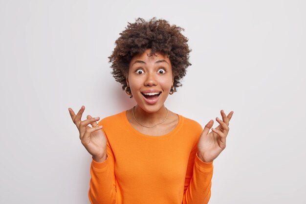 Surprised cheerful Afro American woman stares with great wonder raises hands cannot believe in shocking news wears casual orange jumper isolated over grey wall
