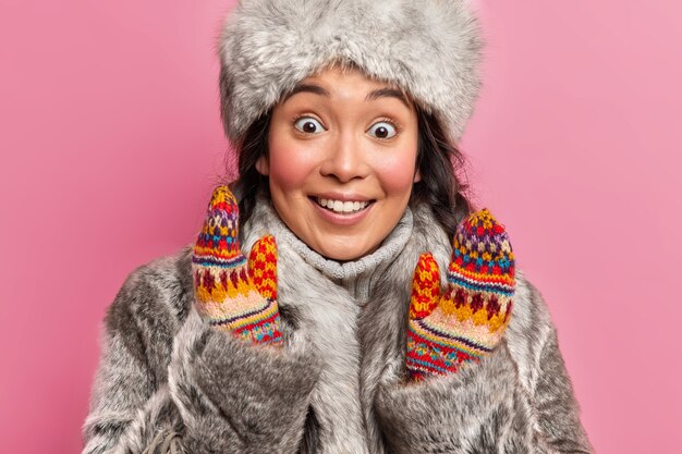 Удивленная жизнерадостная эскимосская девушка смотрит вперед, улыбается, широко поднимает руки, одетая в традиционную серую шубу и шляпу, изолированную над розовой стеной