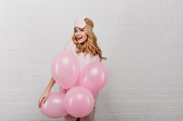 Surprised cheerful girl in sleepmask standing on white wall with party balloons. Portrait of beautiful young lady celebrating something in weekend morning.