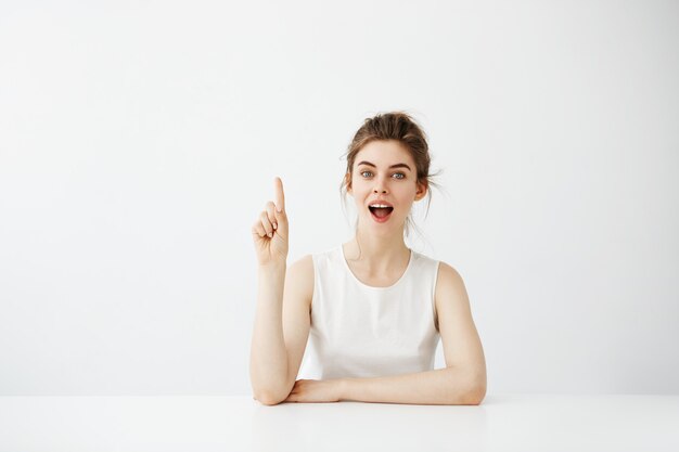 Surprised cheerful pretty woman  with opened mouth pointing finger up sitting at table over white background.