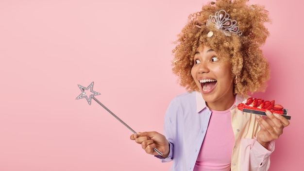 Free photo surprised cheerful woman holds magic wand and delicious strawberry cake exclaims loudly wears crown and shirt isolated over pink background empty space for your advertisement celebration concept