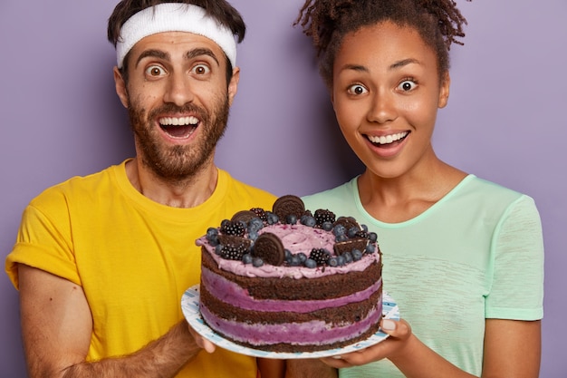 Surprised cheerful woman and man holds big delicious cake on plate, congratulate friend with birthday