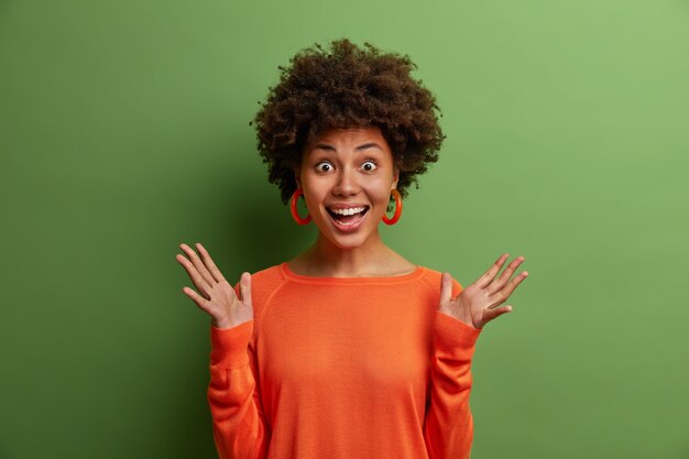 surprised cheerful woman with curly hair raises hands and feels impressed, reacts on awesome surprise prepared by boyfriend, wears orange jumper, isolated on green wall.