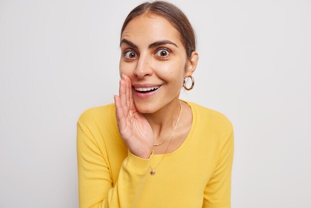 Surprised cheerful woman with dark hair keeps palm near mouth whispers something looks with wondered mysterious expression dressed in yellow jumper isolated over white background. Listen what I know