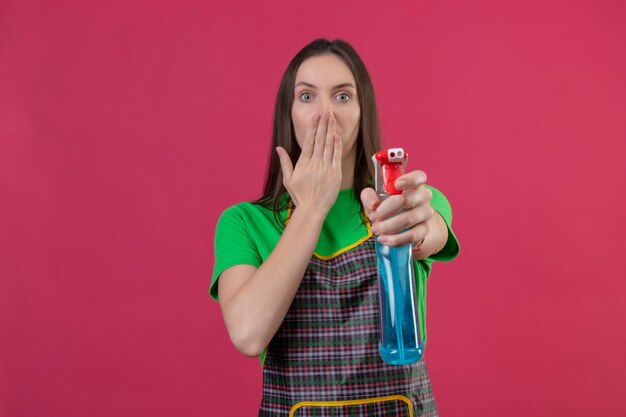 Foto gratuita ragazza giovane di pulizia sorpresa che indossa l'uniforme in guanti che tengono fuori lo spruzzo di pulizia alla bocca coperta della macchina fotografica su fondo rosa isolato