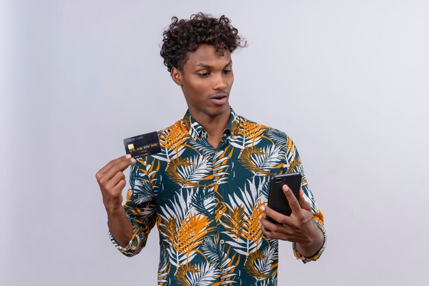 Surprised and confused good-looking dark-skinned man with curly hair showing credit card while looking at mobile phone on a white background