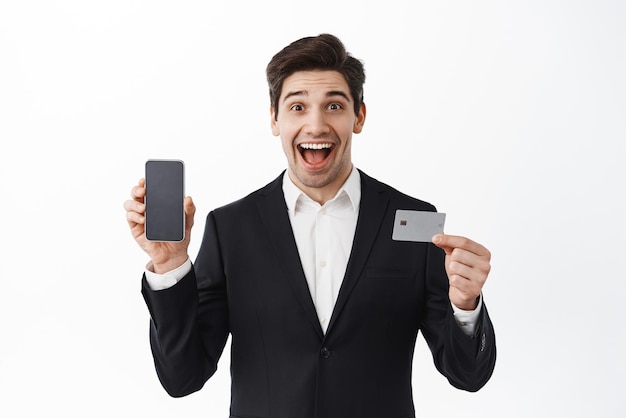 Surprised corporate man businessman in suit shows empty phone screen and credit card smiling amazed new bank app standing over white background