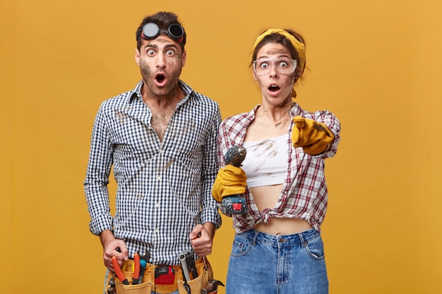 Free photo surprised couple of male and female electrical technicians in protective goggles and overalls having astonished looks, girl with drill pointing index finger, showing something shocking