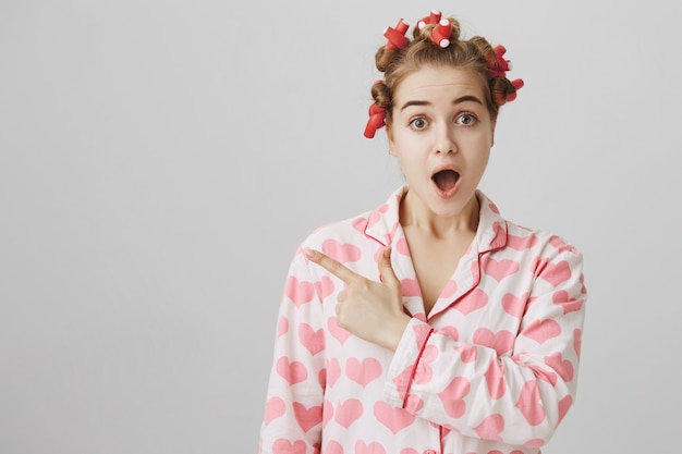 Surprised and curious cute girl in nightwear and hair curlers pointing finger left at banner