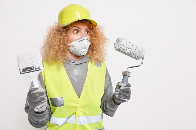 Surprised curly haired woman builder wears construction helmet safety clothe protective mask holds building equipment for house refurbishment stands against white wall copy space for inscription