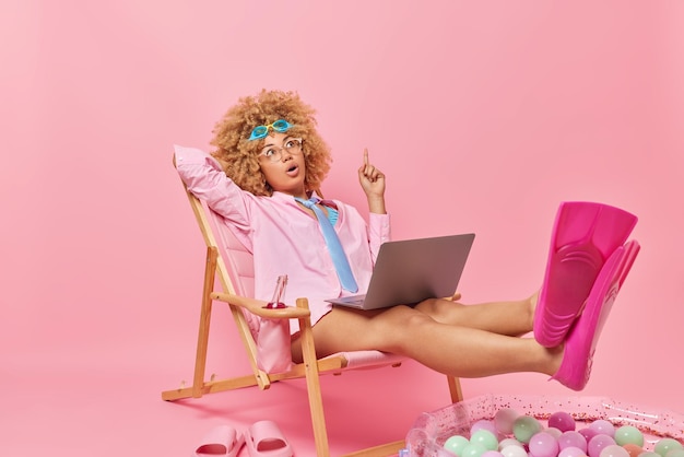 곱슬머리 여성이 셔츠 넥타이와 오리발을 착용하고 긴 의자 라운지에서 노트북 포즈를 사용하는 소셜 네트워크의 여름 방학 블로그에 대한 손가락 오버헤드 꿈에 대해 집중적으로 놀란 곱슬머리 여성