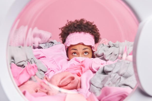 Surprised curly haired woman covered with big stack of laundry focused above poses in washing machine busy washing dirty clothing does domestic chores