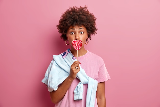 Foto gratuita la donna dai capelli ricci sorpresa copre la bocca con un gustoso lecca-lecca di caramelle a forma di cuore, andando a fare una passeggiata all'aperto con il fidanzato, vestito con una maglietta casual e un maglione legato sulla spalla. persone, giovani