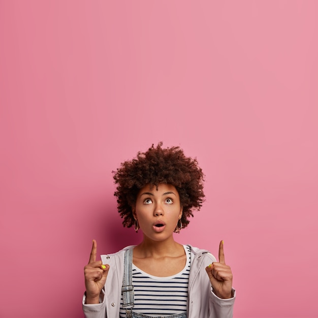 Foto gratuita la donna sorpresa dai capelli ricci si sente imbarazzata, punta il dito indice verso l'alto, mostra un promo straordinario, vestita in modo casual, racconta di un evento interessante che si svolge nelle vicinanze al piano di sopra, apre la bocca per lo stupore