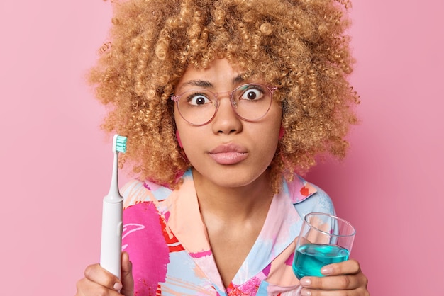 Free photo surprised curly haired woman going to brush teeth holds electric toothbrush and glass of mouthwash wears spectacles for good sight wears casual shirt isolated over pink background daily hygiene