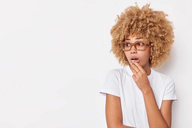 Surprised curly haired woman keeps mouth opened focused away stares impressed at something shocking wears casual t shirt and spectacles isolated over white background copy space away for text