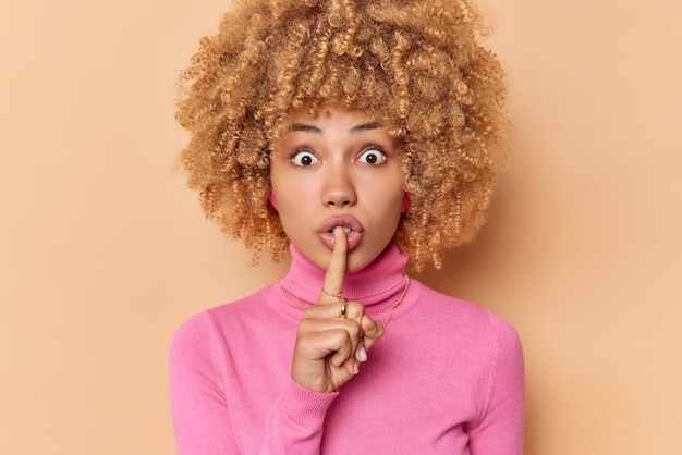 Free photo surprised curly haired woman presses index finger over lips asks to keep promise has shocked expression makes silence gesture dressed in casual pink turtleneck isolated over beige background