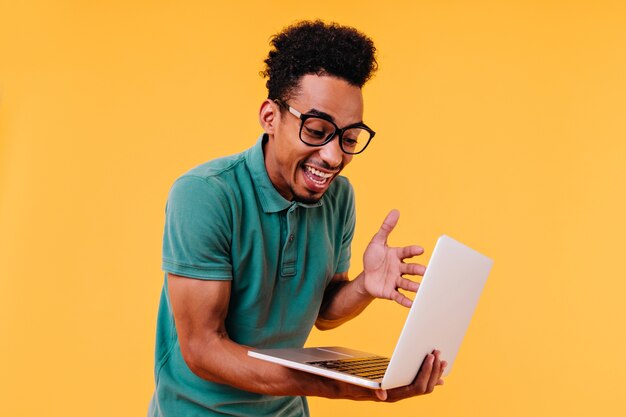 surprised curly male student looking at laptop screen. indoor shot of african freelancer wears glasses.